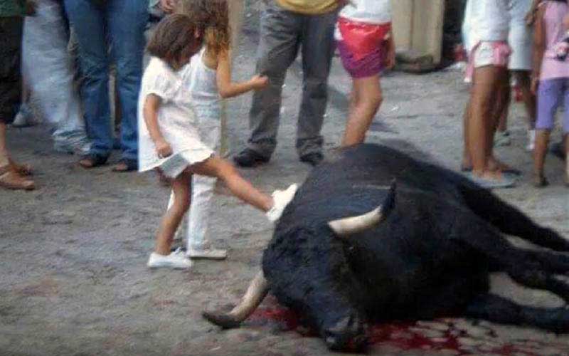 Une jeune fille donne un coup de pied à la tête d'un taureau mort au sol lors d'une rue ou d'une place publique, entourée de spectateurs.